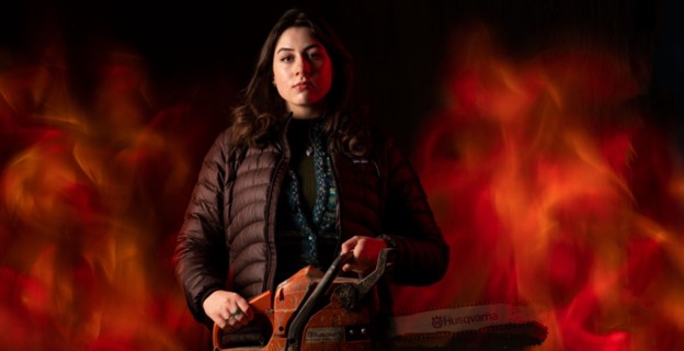 Pitt student Helene Tracey wields a chainsaw, staring into the camera against a flaming background.